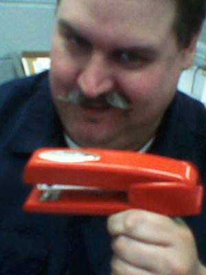 Headshot of a person with a mustache, holding a red stapler up in front of them